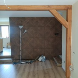 URB Krzysztof Kochanowski - Pokój z drewnianą konstrukcją, panelami ściennymi o geometrycznym wzorze i odkurzaczem na podłodze z paneli, widoczne niedokończone prace elektryczne.