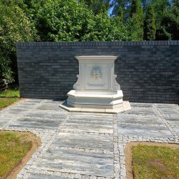 URB Krzysztof Kochanowski - Ogród z fontanną ozdobioną motywem lwa, otoczoną szarą kostką brukową ułożoną w kwadraty i obramowaną drobną kostką, na tle ciemnego muru z cegły.