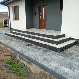 URB Krzysztof Kochanowski - Nowoczesne wejście do domu z kostki brukowej w odcieniach szarości, z trzema stopniami wykończonymi białymi płytami, widoczne świeżo ułożone obrzeża i fragment trawnika z przewodami.