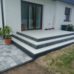 URB Krzysztof Kochanowski - Nowoczesne schody wejściowe z kostki brukowej, wykończone szarymi obrzeżami, prowadzące do białego domu z brązowymi oknami, obok donica z rośliną i fragment ułożonej kostki na ziemi.