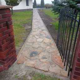 URB Krzysztof Kochanowski - Ścieżka z nieregularnych kamieni prowadząca od bramki do domu, widoczna z perspektywy bramki, z ceglanymi słupkami po bokach.