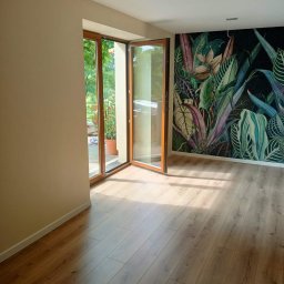 URB Krzysztof Kochanowski - Jasne panele podłogowe ułożone w pokoju z otwartymi drewnianymi drzwiami balkonowymi i ścianą pokrytą tapetą w motywy roślinne.