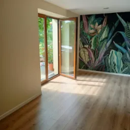 Jasne panele podłogowe ułożone w pokoju z otwartymi drewnianymi drzwiami balkonowymi i ścianą pokrytą tapetą w motywy roślinne.