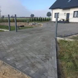 Nowo ułożona, szara kostka brukowa tworząca podjazd do domu jednorodzinnego, widok z poziomu gruntu, z widoczną linią granicy z piaskiem i trawnikiem.