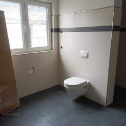 URB Krzysztof Kochanowski - Wnętrze łazienki w trakcie remontu: szare płytki podłogowe, białe płytki ścienne z ciemnoszarym pasem dekoracyjnym, wisząca toaleta, okno z widokiem na rusztowanie, karton zabezpieczający w rogu.