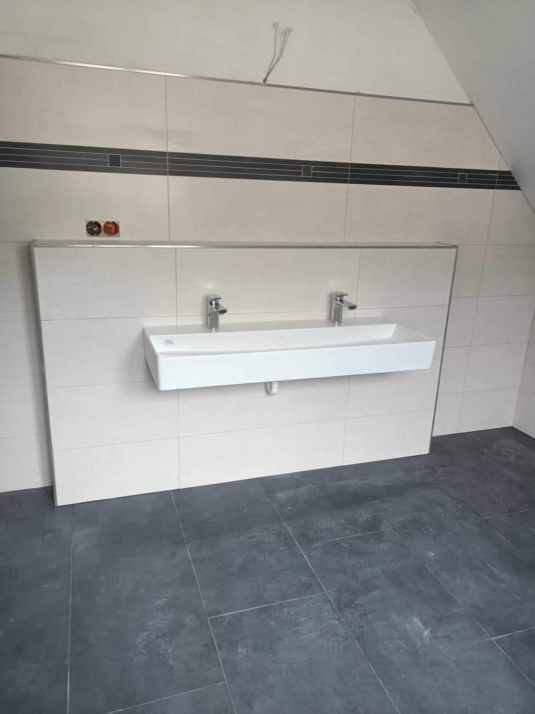 Wykończona łazienka na poddaszu: białe prostokątne płytki ścienne z poziomym pasem ciemnych płytek, podwójna umywalka nablatowa z chromowanymi bateriami, szare płytki podłogowe, widoczne wystające...