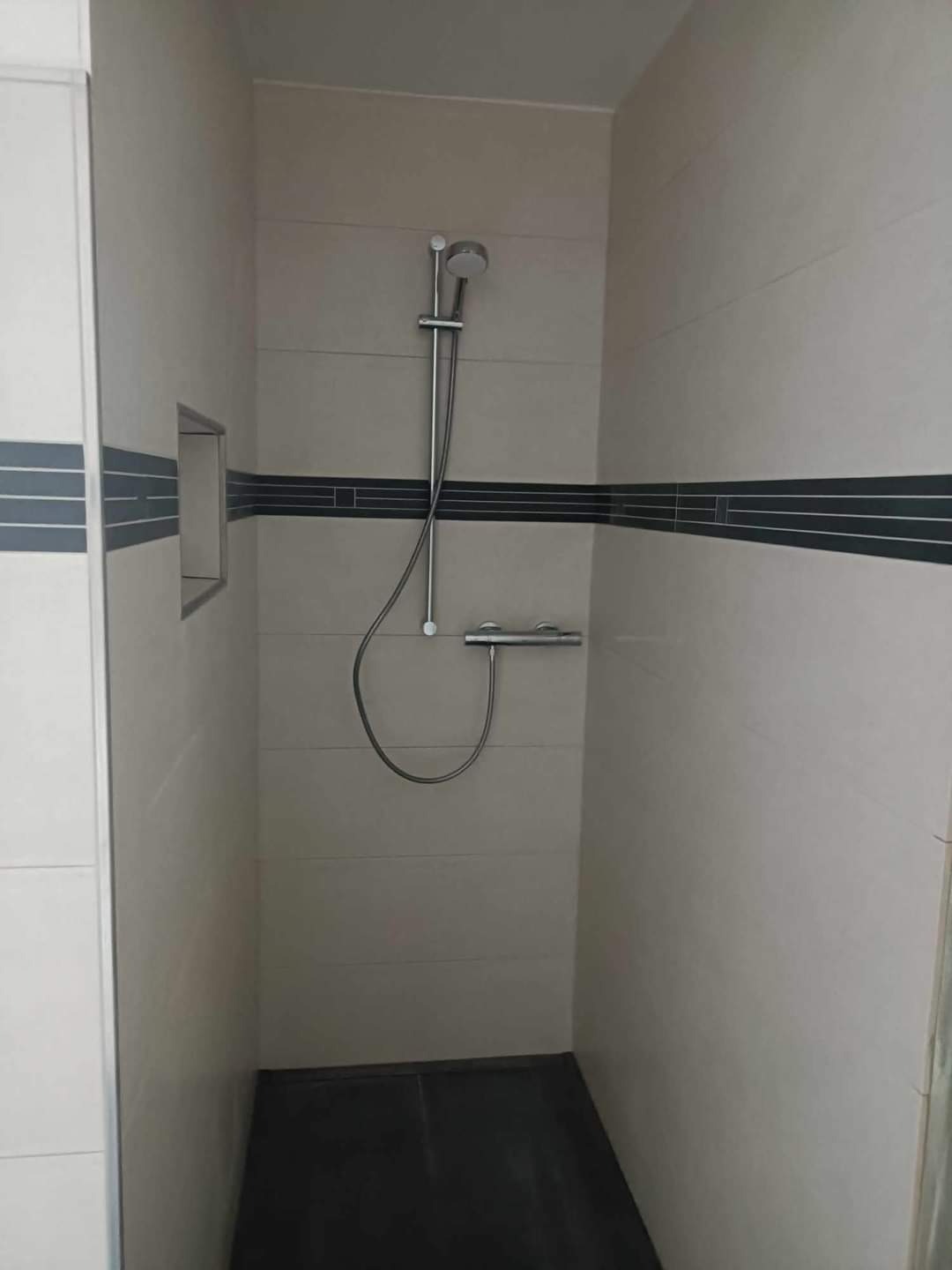 Wykończona kabina prysznicowa z beżowymi płytkami z ciemnym paskiem dekoracyjnym i czarną podłogą, widok z góry.