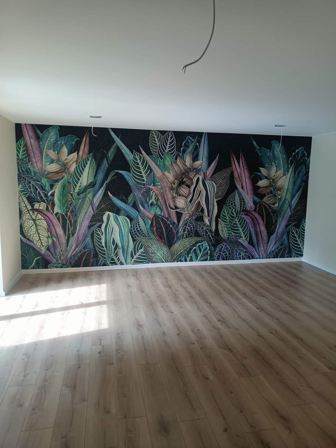 Pokój po remoncie z drewnopodobną podłogą i fototapetą z motywem tropikalnych liści na czarnym tle, widoczne kable wystające z sufitu.