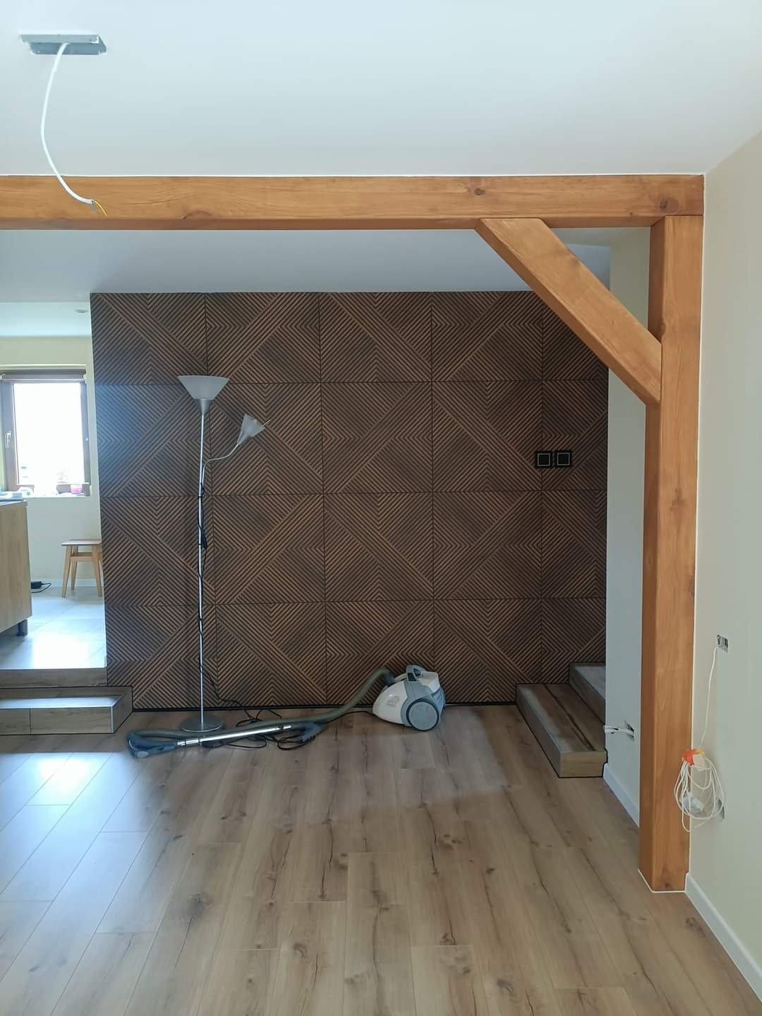 Pokój z drewnianą konstrukcją, panelami ściennymi o geometrycznym wzorze i odkurzaczem na podłodze z paneli, widoczne niedokończone prace elektryczne.