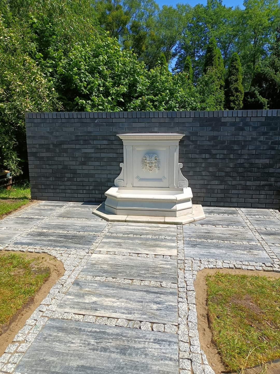 Ogród z fontanną ozdobioną motywem lwa, otoczoną szarą kostką brukową ułożoną w kwadraty i obramowaną drobną kostką, na tle ciemnego muru z cegły.