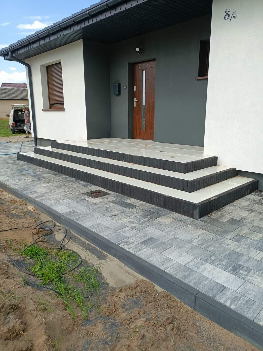 Nowoczesne wejście do domu z kostki brukowej w odcieniach szarości, z trzema stopniami wykończonymi białymi płytami, widoczne świeżo ułożone obrzeża i fragment trawnika z przewodami.