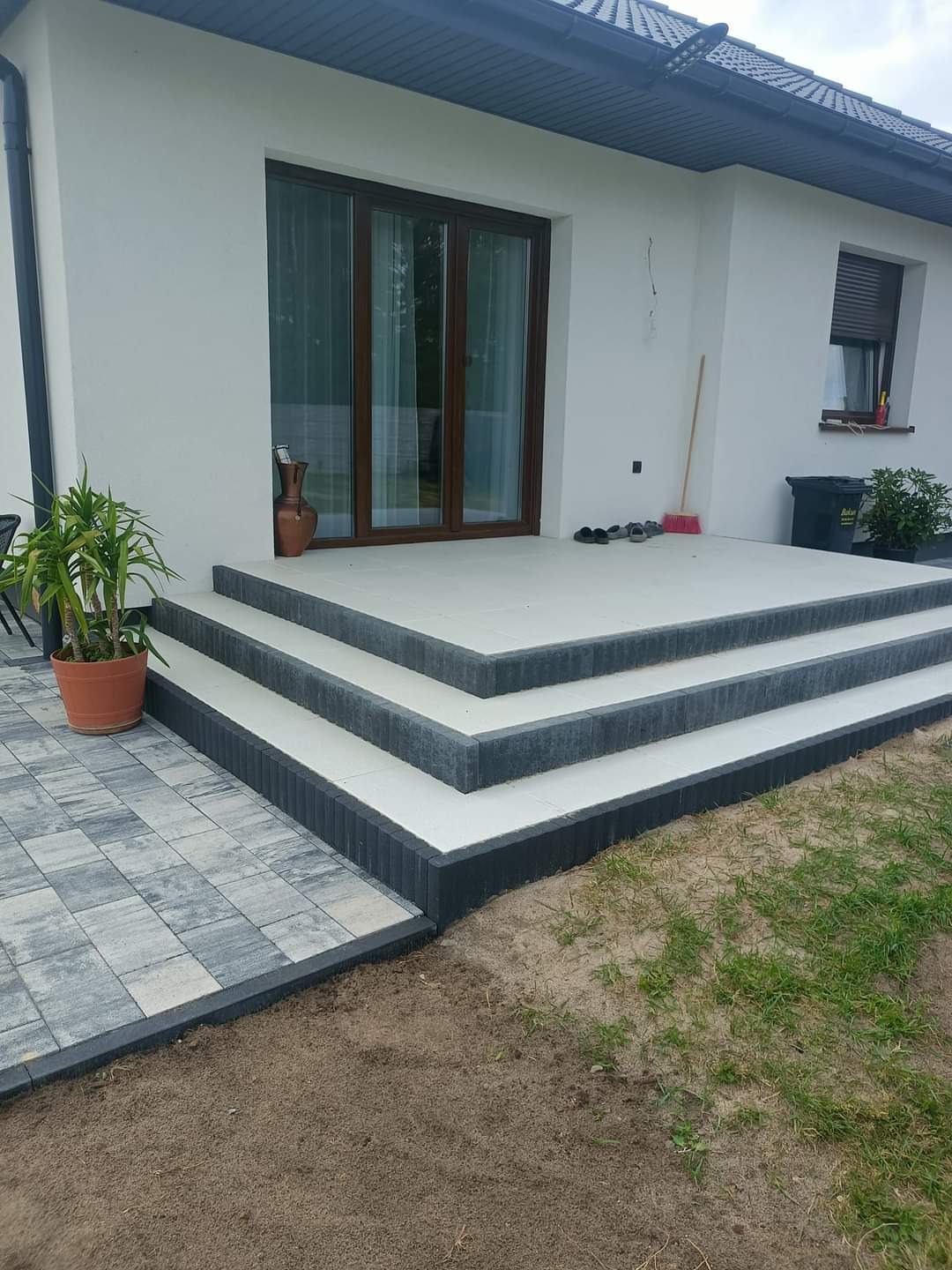 Nowoczesne schody wejściowe z kostki brukowej, wykończone szarymi obrzeżami, prowadzące do białego domu z brązowymi oknami, obok donica z rośliną i fragment ułożonej kostki na ziemi.