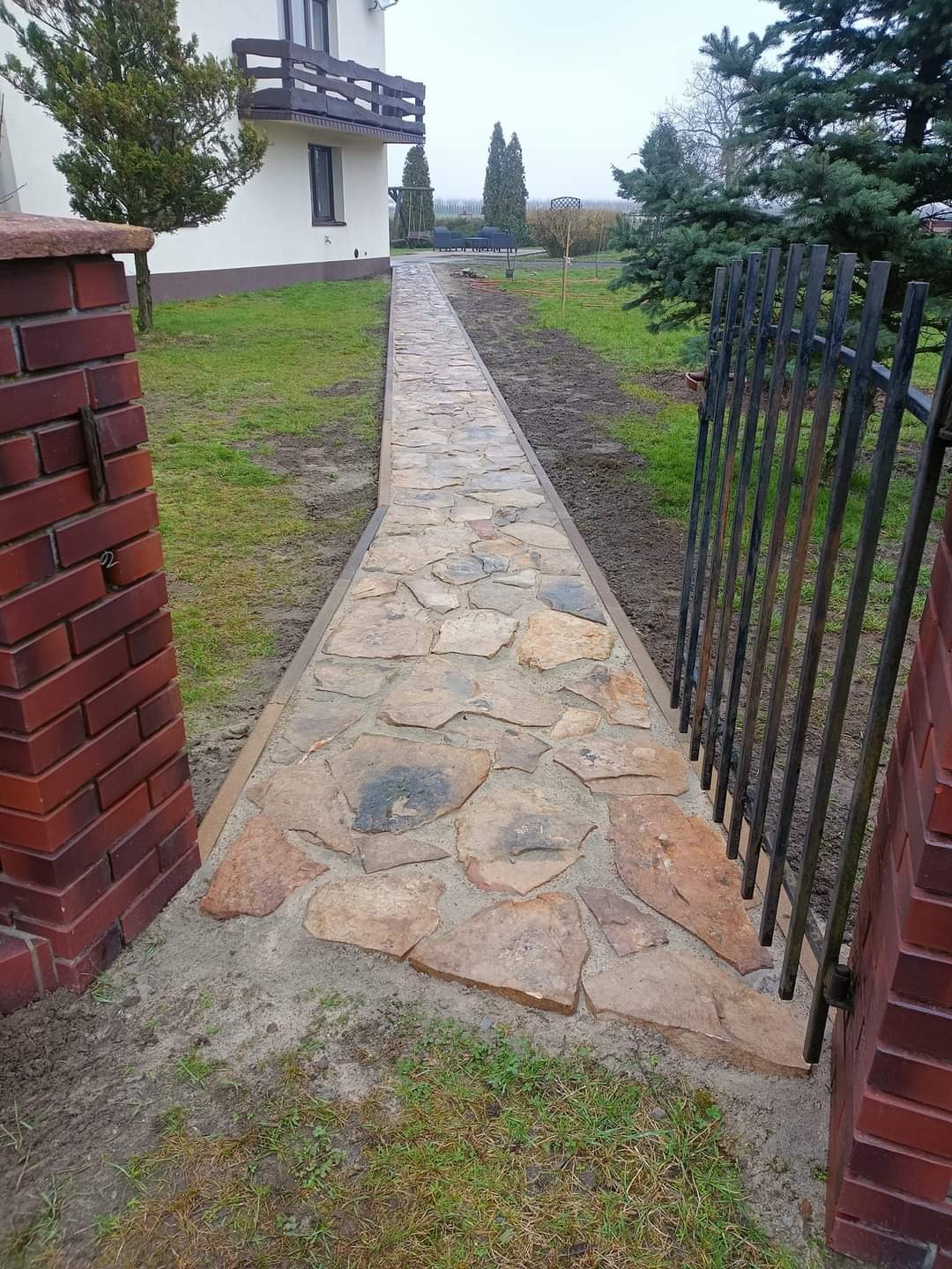 Ścieżka z nieregularnych kamieni prowadząca od bramki do domu, widoczna z perspektywy bramki, z ceglanymi słupkami po bokach.