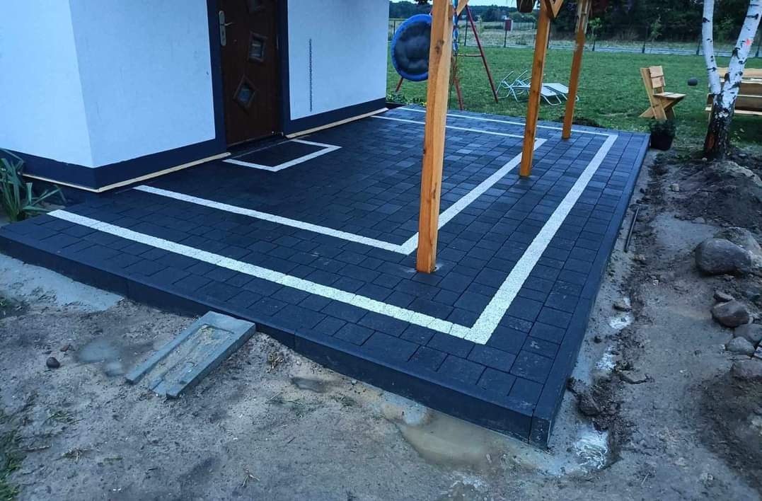 Nowoczesny taras z ciemnej kostki brukowej obramowany białymi pasami, z drewnianymi słupami zadaszenia i widokiem na trawnik z huśtawką w tle.