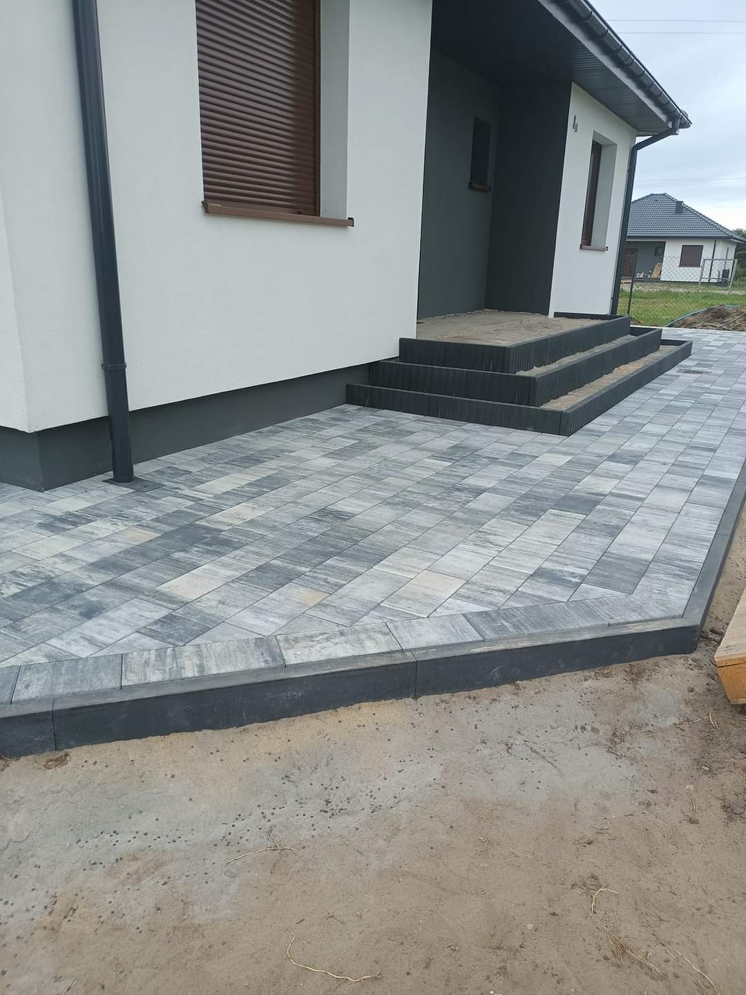 Ułożona kostka brukowa w odcieniach szarości przed domem z trzema betonowymi schodami i brązową roletą okienną, widoczne obrzeża i fragment niezakończonej nawierzchni.