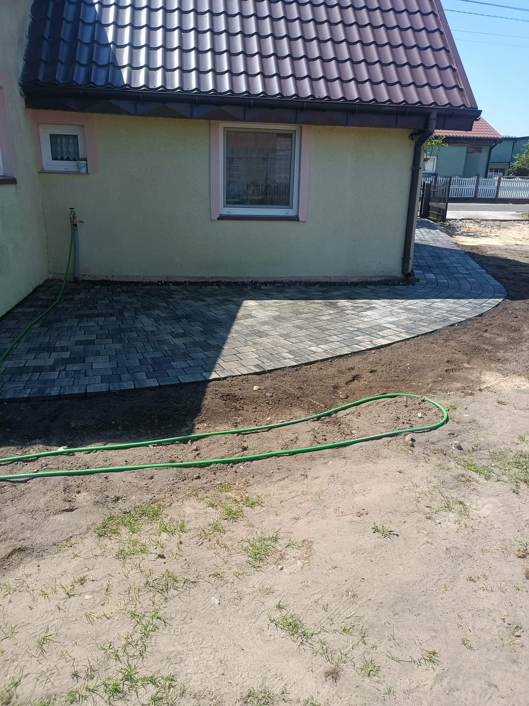 Nowa, szara kostka brukowa ułożona przy ścianie domu z zieloną elewacją i brązowym dachem, widoczny zielony wąż ogrodowy na piaszczystym podłożu.