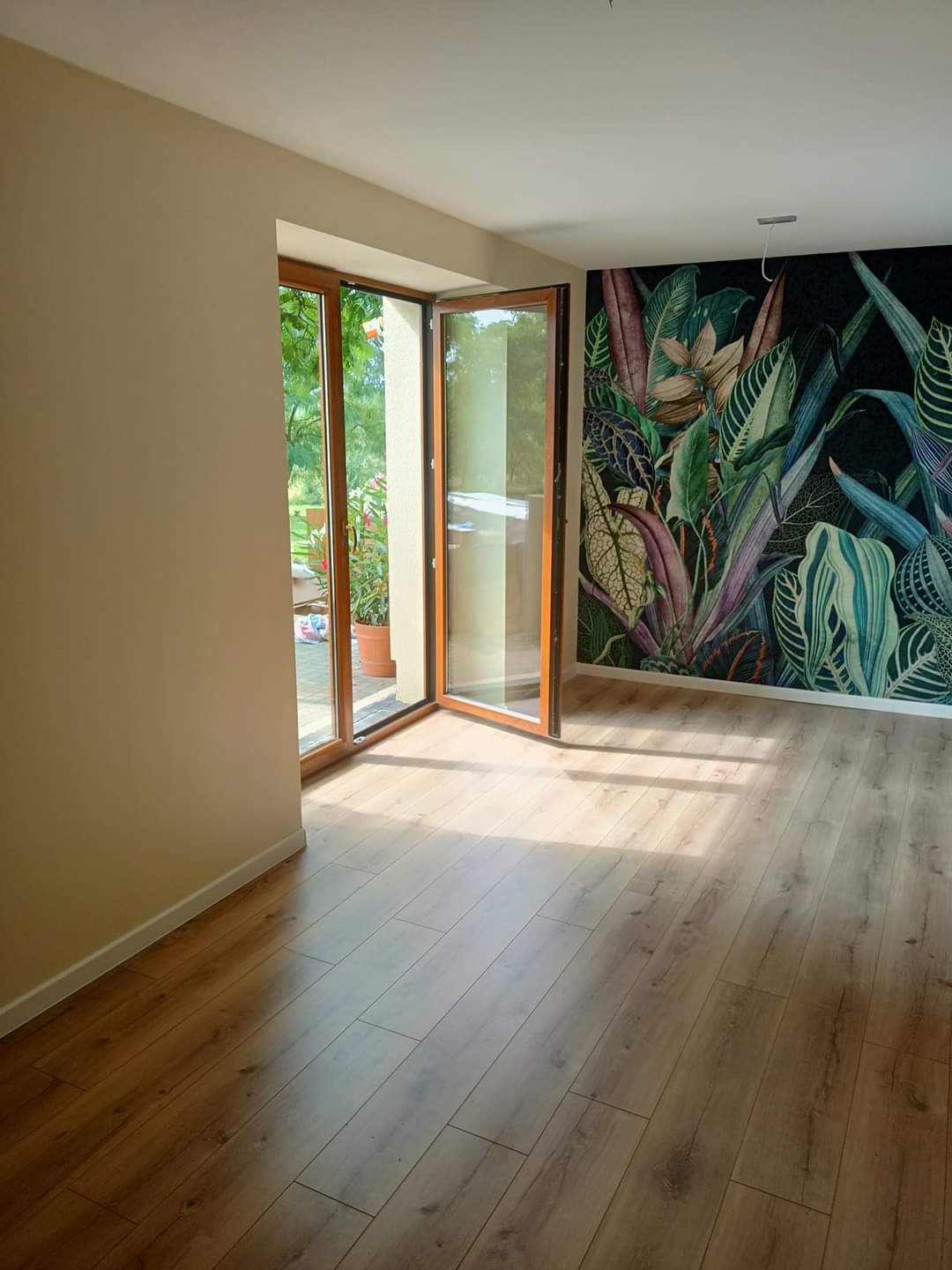 Jasne panele podłogowe ułożone w pokoju z otwartymi drewnianymi drzwiami balkonowymi i ścianą pokrytą tapetą w motywy roślinne.