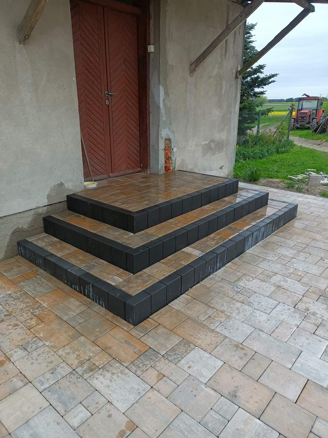 Nowo ułożone, trójstopniowe schody z kostki brukowej prowadzące do wejścia budynku, z widocznym traktorem w tle.