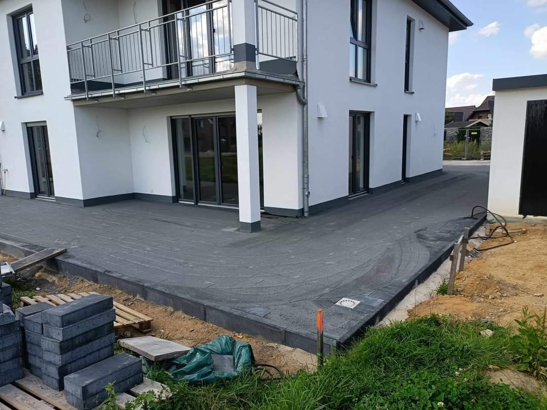 Nowo ułożona, wilgotna, szara kostka brukowa na podjeździe przed nowoczesnym, białym domem z balkonem, widoczne palety z kostką i trawa.