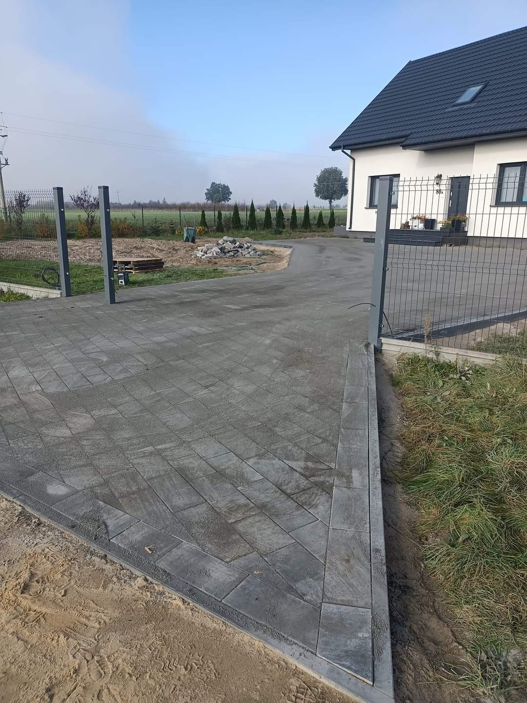 Nowo ułożona, szara kostka brukowa tworząca podjazd do domu jednorodzinnego, widok z poziomu gruntu, z widoczną linią granicy z piaskiem i trawnikiem.