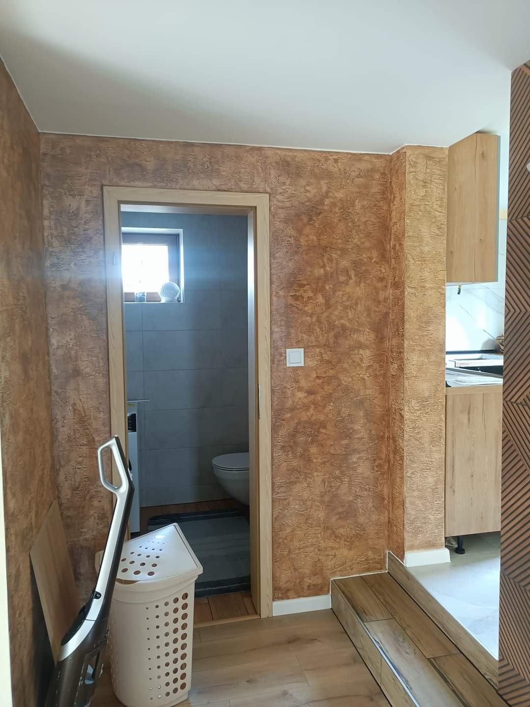 Wnętrze mieszkania po nałożeniu dekoracyjnej, brązowej farby strukturalnej na ścianach, widoczne wejście do łazienki i fragment zabudowy kuchennej.