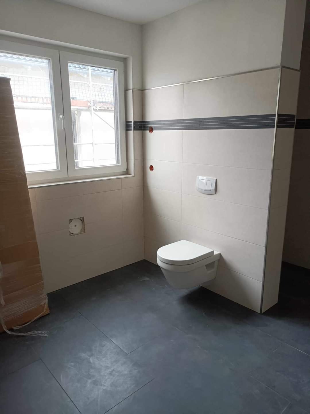 Wnętrze łazienki w trakcie remontu: szare płytki podłogowe, białe płytki ścienne z ciemnoszarym pasem dekoracyjnym, wisząca toaleta, okno z widokiem na rusztowanie, karton zabezpieczający w rogu.