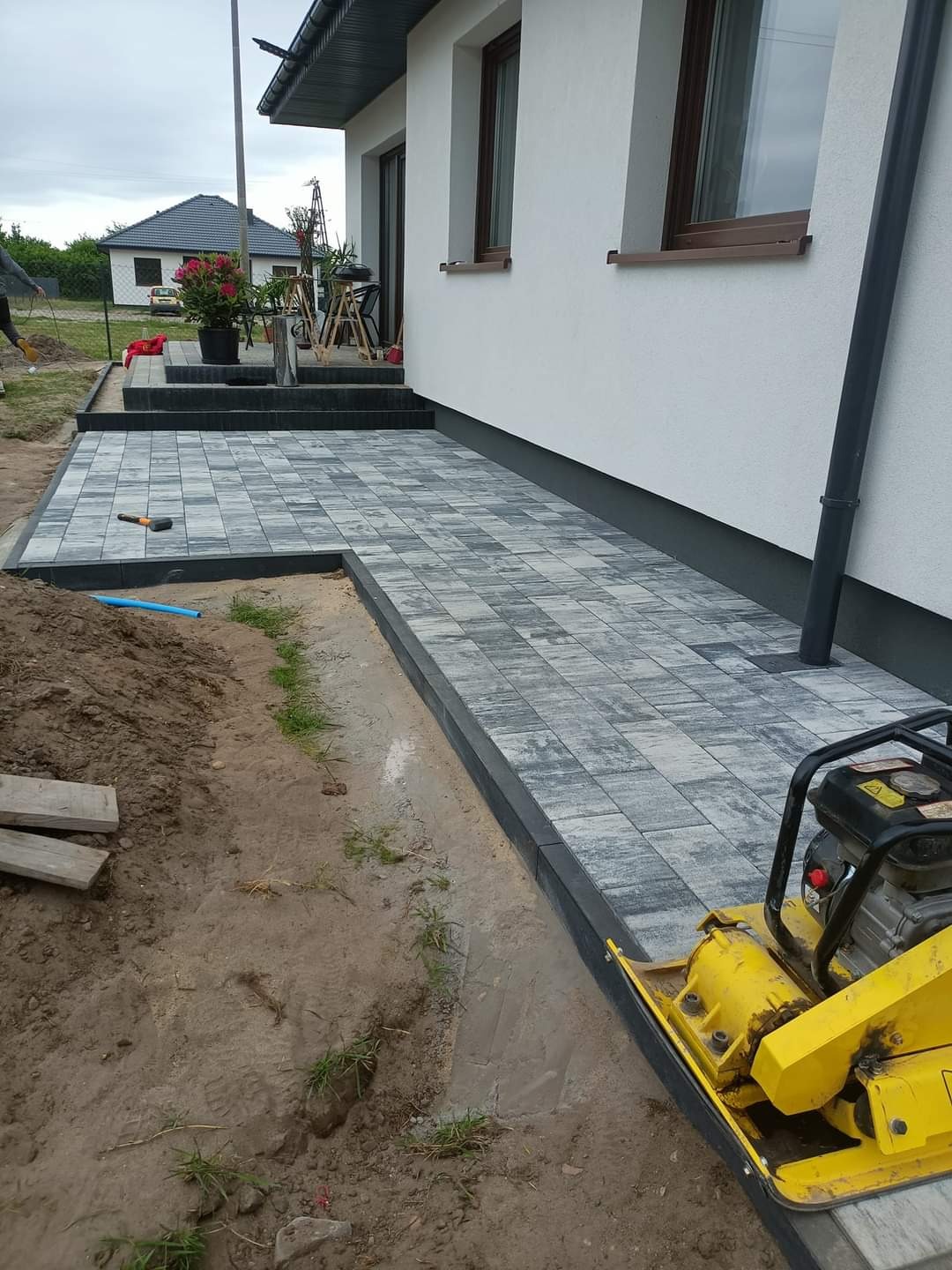 Układanie szarej kostki brukowej przy domu z użyciem zagęszczarki w trakcie prac ziemnych, widoczne obrzeża i przygotowane podłoże.