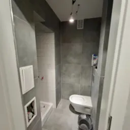 Wnętrze łazienki w trakcie remontu: szare płytki wielkoformatowe na ścianach, białe płytki ułożone w jodełkę w kabinie prysznicowej, wisząca toaleta, wiadro z zaprawą i niedokończone oświetlenie...