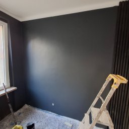 ykonczenia wnetrz maksym merhes - Świeżo pomalowane na ciemny grafit ściany w pokoju, zabezpieczona folią podłoga, drabina malarska i akcesoria malarskie widoczne w rogu, dekoracyjna ściana z pionowych paneli.