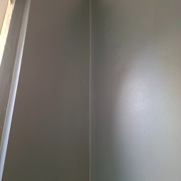 ykonczenia wnetrz maksym merhes - Świeżo pomalowany narożnik pokoju w odcieniach szarości, widoczne niedoskonałości na ścianie przy suficie.