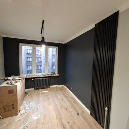 ykonczenia wnetrz maksym merhes - Pokój po remoncie: ciemne ściany z drewnianymi lamelami, jasna podłoga z paneli, okno z widokiem na budynek, karton z narzędziami i resztki materiałów na podłodze.