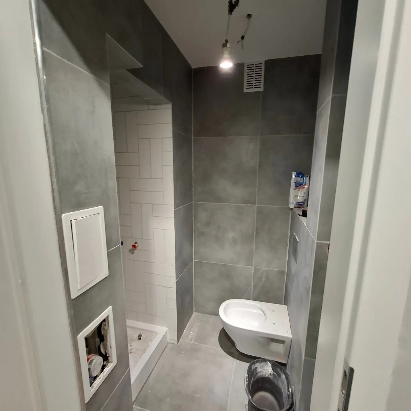 Szare płytki wielkoformatowe w łazience z widoczną kabiną prysznicową wyłożoną białą cegiełką, częściowo zamontowana toaleta i wiadro z resztkami zaprawy.