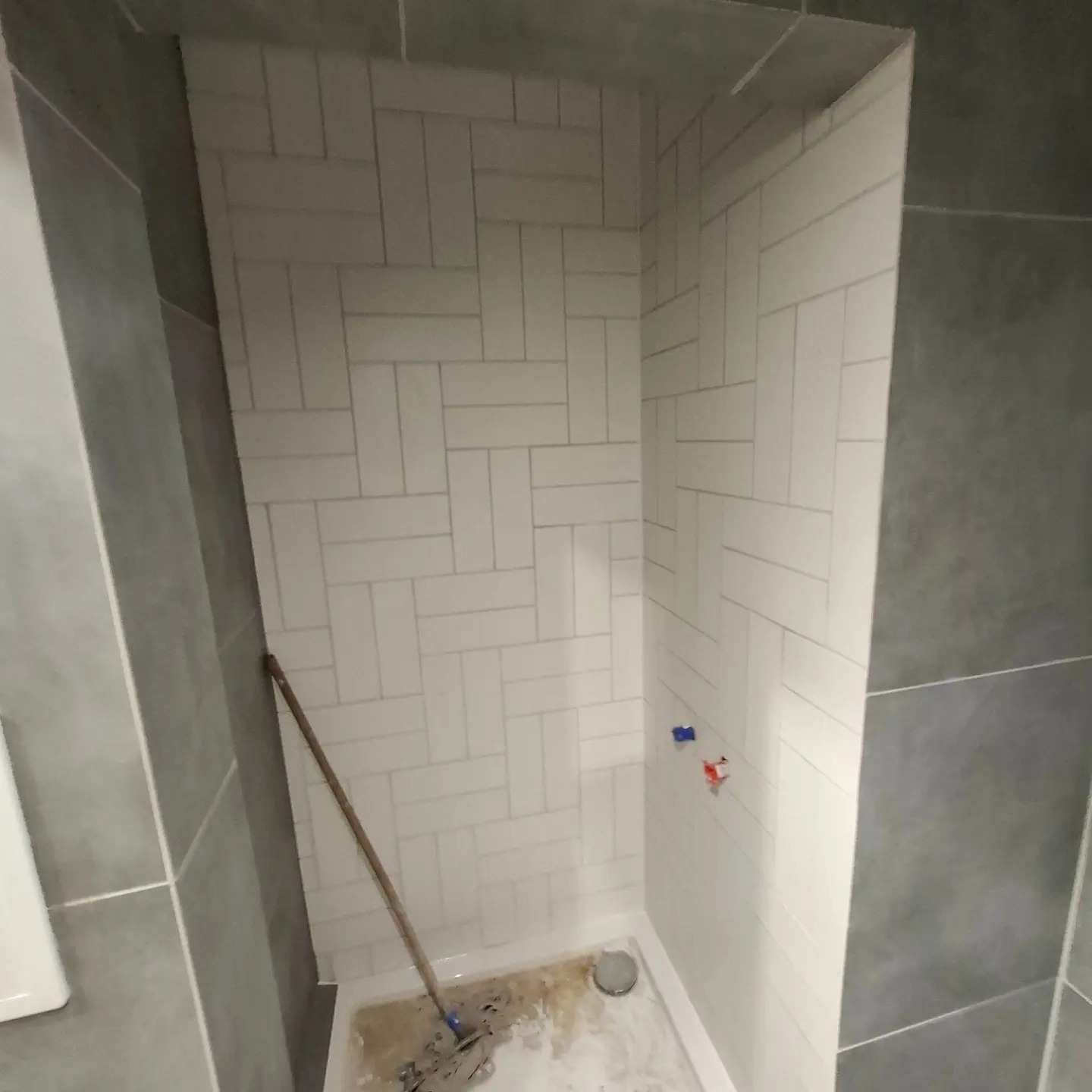 Wnęka prysznicowa w trakcie wykańczania, białe płytki ułożone w jodełkę na ścianach, widoczne przyłącza wodne, na podłodze resztki zaprawy i narzędzie.