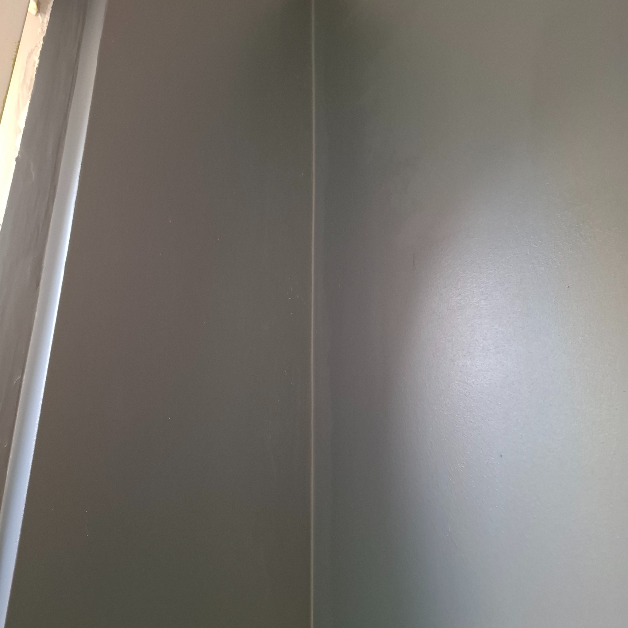 Świeżo pomalowany narożnik pokoju w odcieniach szarości, widoczne niedoskonałości na ścianie przy suficie.