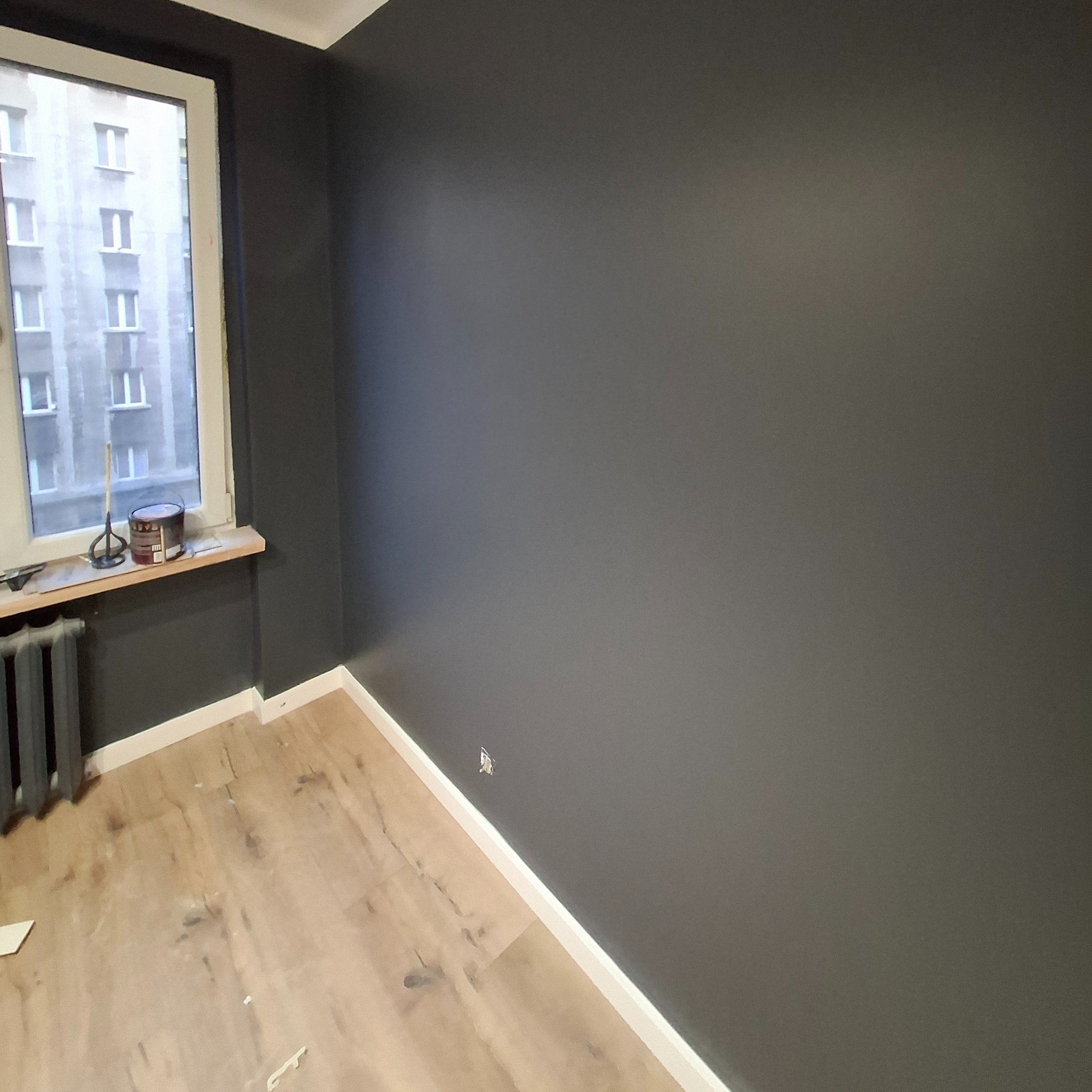 Świeżo pomalowany na szaro pokój z widocznymi śladami pracy: puszka farby i mieszadło na parapecie, jasne panele podłogowe z drobnymi zabrudzeniami, białe listwy przypodłogowe.