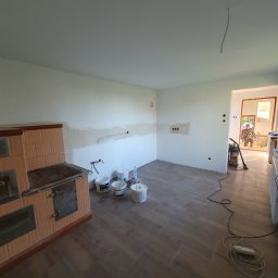 BRO-BUILD - Wnętrze kuchni w trakcie remontu: widoczne szpachlowane fragmenty ścian, piec kaflowy, wiaderka z farbą, odkurzacz i płytki podłogowe imitujące drewno.