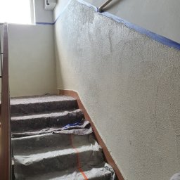 BRO-BUILD - Klatka schodowa w trakcie prac wykończeniowych: świeżo nałożony tynk na ścianie, zabezpieczone schody folią i materiałem ochronnym, widoczny fragment listwy przypodłogowej i taśmy malarskiej.