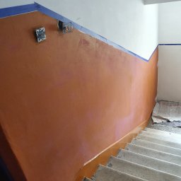 BRO-BUILD - Malowanie klatki schodowej w Lubaczowie, widoczne zabezpieczone taśmą malarską krawędzie, odsłonięte puszki instalacyjne i świeżo pomalowana ściana w odcieniach brązu.