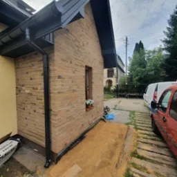 Fragment domu z elewacją z cegły klinkierowej, widoczny system rynnowy w kolorze antracytowym, okno z kwiatami w doniczce, teren wok&oacute;ł domu przygotowany pod prace ziemne.