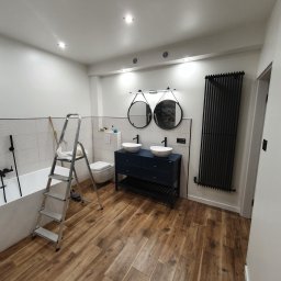 BRO-BUILD - Nowoczesna łazienka z drewnopodobną podłogą, podwójną umywalką nablatową na granatowej szafce, czarnym grzejnikiem drabinkowym i drabiną opartą o wannę przy ścianie z mozaiką.