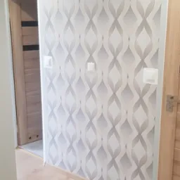 Wnętrze korytarza po remoncie: ściana pokryta tapetą w szaro-białe wzory, jasna drewniana podłoga, drzwi po lewej stronie z ciemniejszymi wstawkami, po prawej stronie jasne drzwi z metalową klamką,...