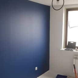 BRO-BUILD - Świeżo pomalowany pokój z jedną ścianą w kolorze granatowym, widoczny sufit, podłoga przygotowana do dalszych prac wykończeniowych, widoczne worki z materiałami budowlanymi i odkurzacz przemysłowy,...