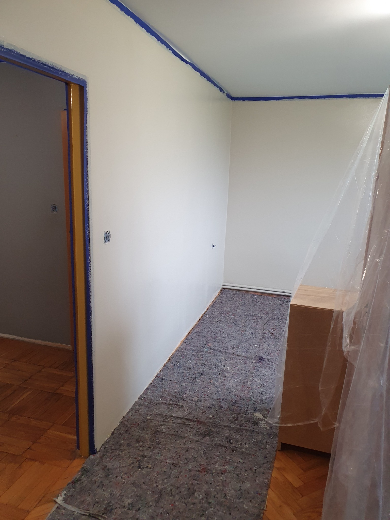 Przygotowane do malowania wnętrze: świeżo pomalowane na biało ściany zabezpieczone niebieską taśmą malarską przy suficie i framugach, podłoga przykryta szarą matą ochronną, meble osłonięte folią...