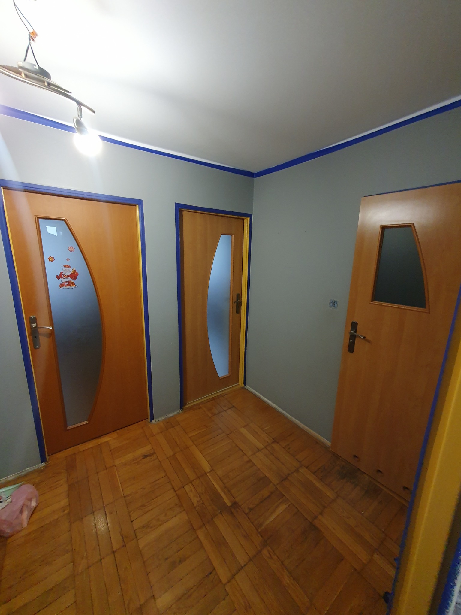 Wnętrze pokoju z trzema drewnianymi drzwiami z szybami o nietypowych kształtach, ściany pomalowane na szaro z niebieskim paskiem przy suficie, parkiet na podłodze.