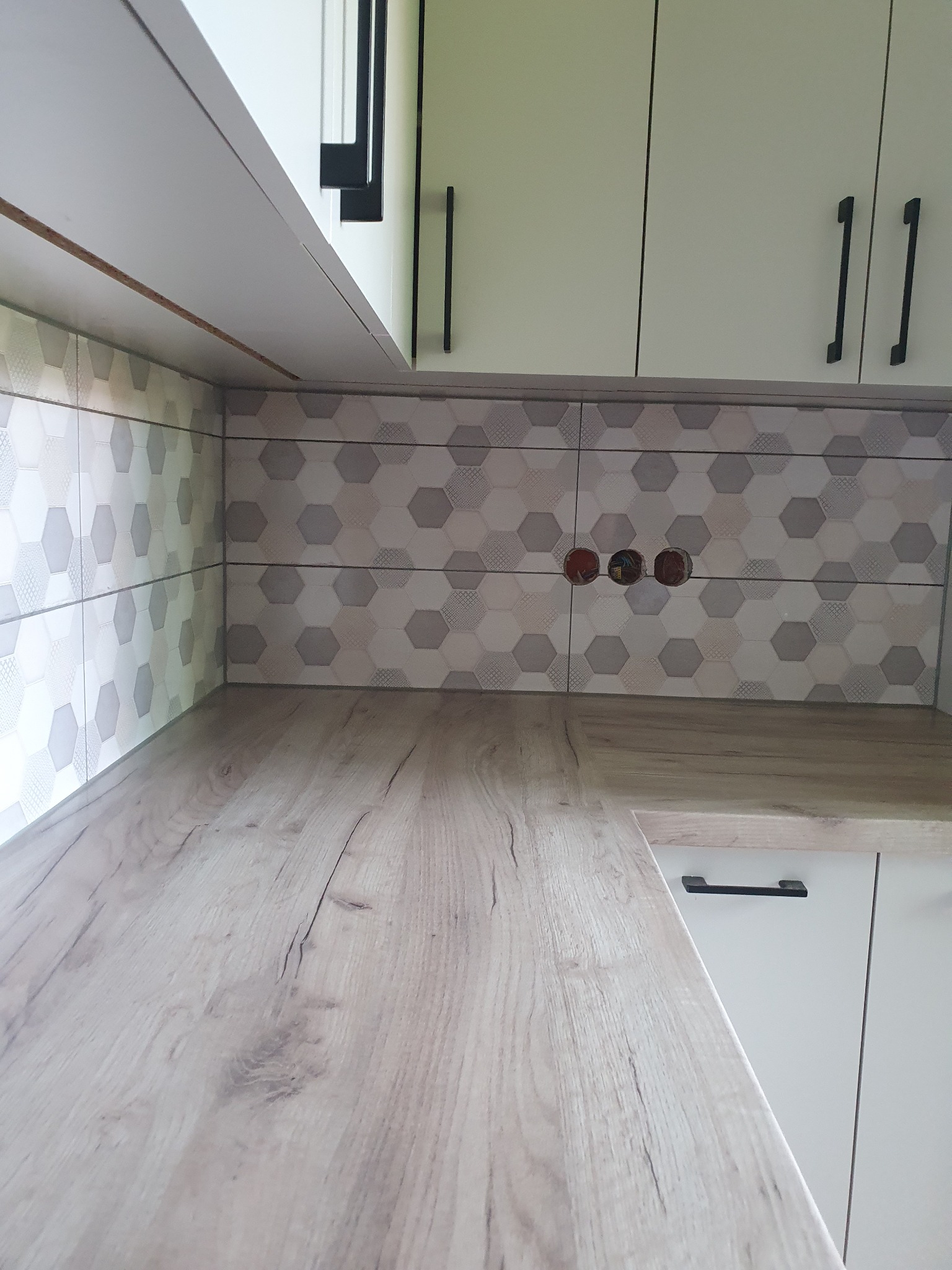 Fragment nowej kuchni z drewnianym blatem, heksagonalnymi płytkami i białymi szafkami z czarnymi uchwytami, widoczne gniazdka elektryczne bez osprzętu.