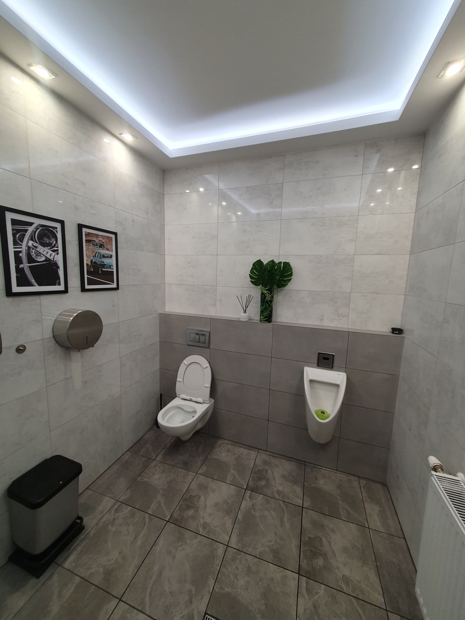 Nowoczesna łazienka publiczna z szarymi płytkami, podświetlanym sufitem, wiszącą toaletą, pisuar z zielonym wkładem, dozownik do papieru i dekoracyjne obrazy w czarnych ramkach.