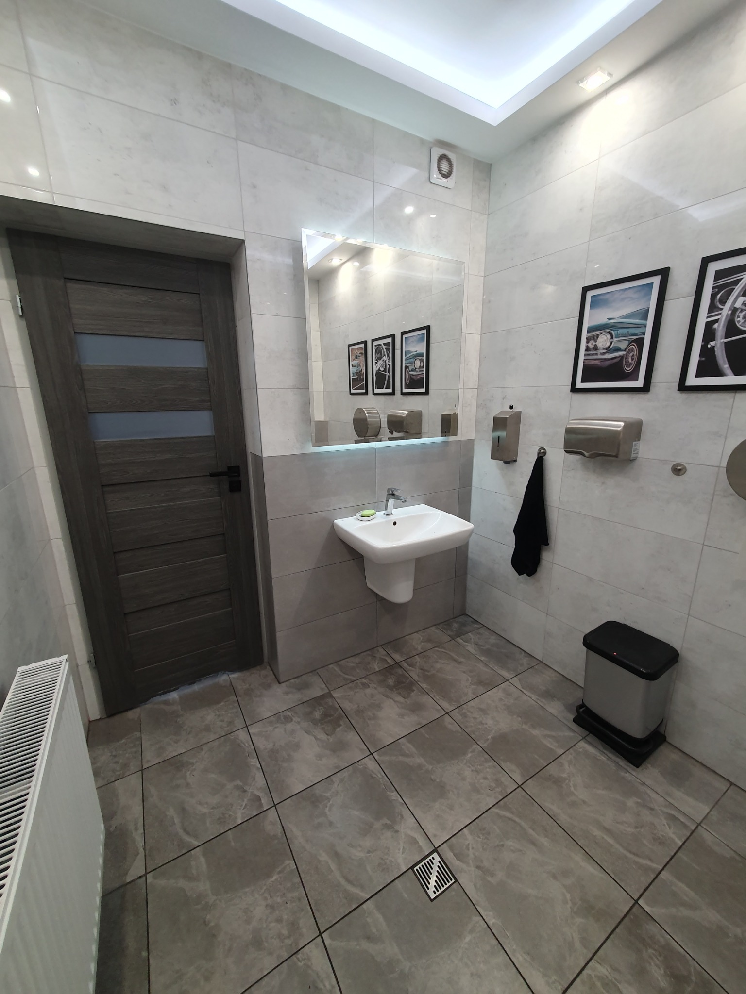 Nowoczesna toaleta publiczna z marmurowymi płytkami, wiszącą umywalką, lustrem z wbudowanym oświetleniem, dozownikiem mydła, suszarką do rąk, czarnym ręcznikiem i dekoracyjnymi obrazami samochodów...