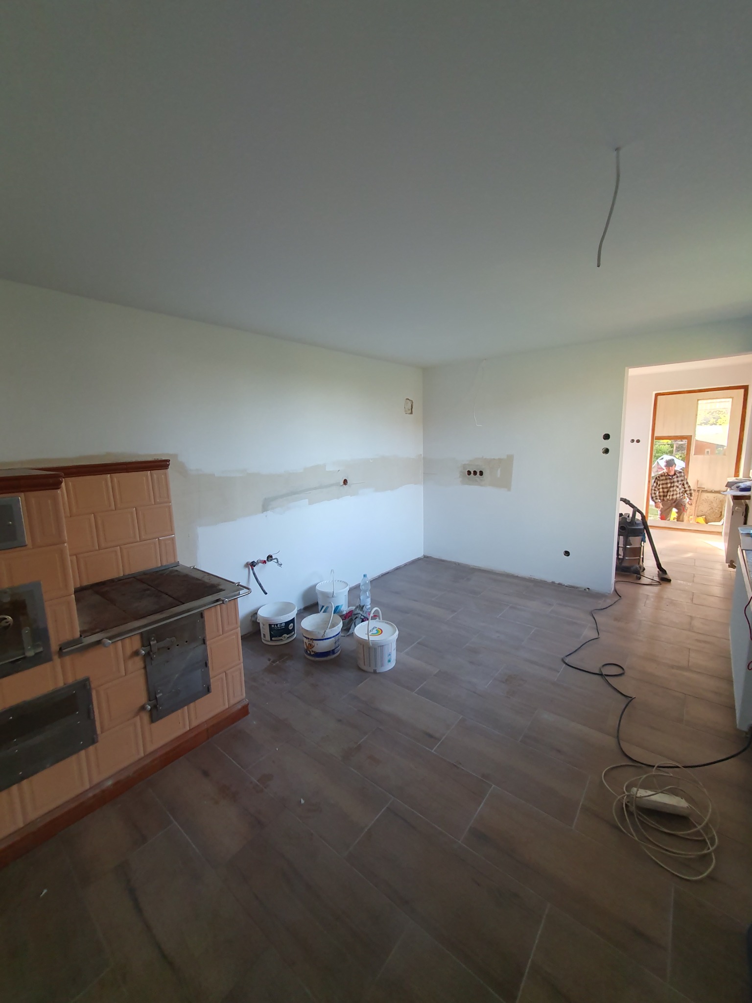 Wnętrze kuchni w trakcie remontu: widoczne szpachlowane fragmenty ścian, piec kaflowy, wiaderka z farbą, odkurzacz i płytki podłogowe imitujące drewno.