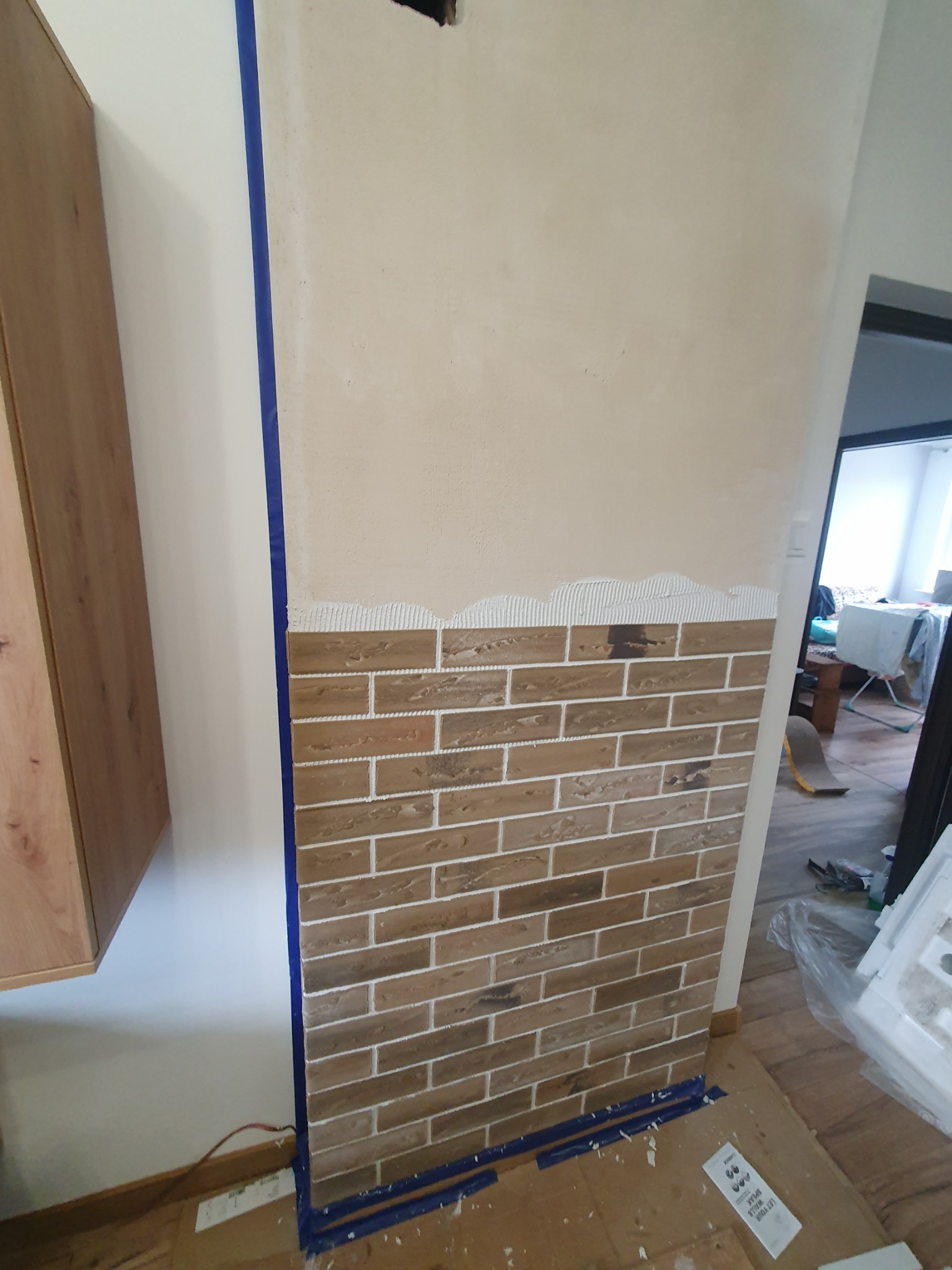 Ściana w trakcie wykańczania z częściowo położoną okładziną cegiełkową, zabezpieczona taśmą malarską, widoczne ślady zaprawy klejowej, reszta ściany przygotowana pod wykończenie.