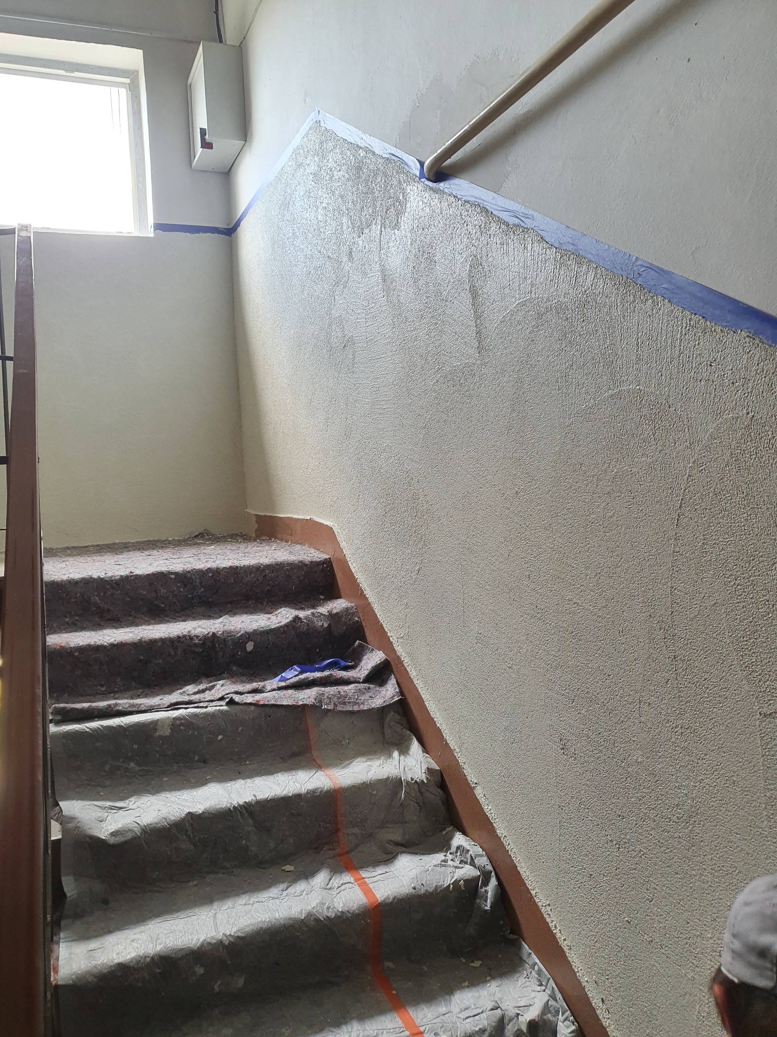 Klatka schodowa w trakcie prac wykończeniowych: świeżo nałożony tynk na ścianie, zabezpieczone schody folią i materiałem ochronnym, widoczny fragment listwy przypodłogowej i taśmy malarskiej.
